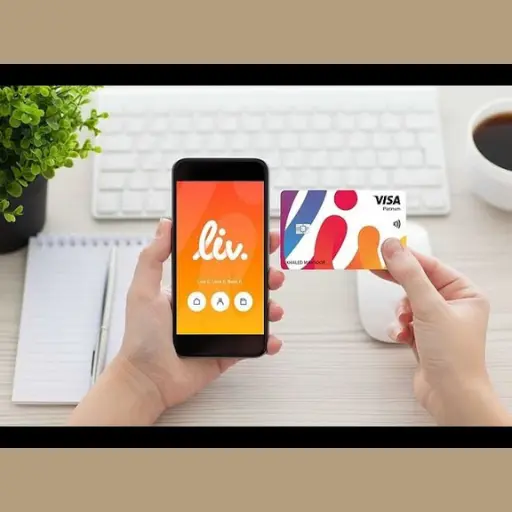 Hand holding a Liv Bank debit card and smartphone showing the Liv app on a desk setup